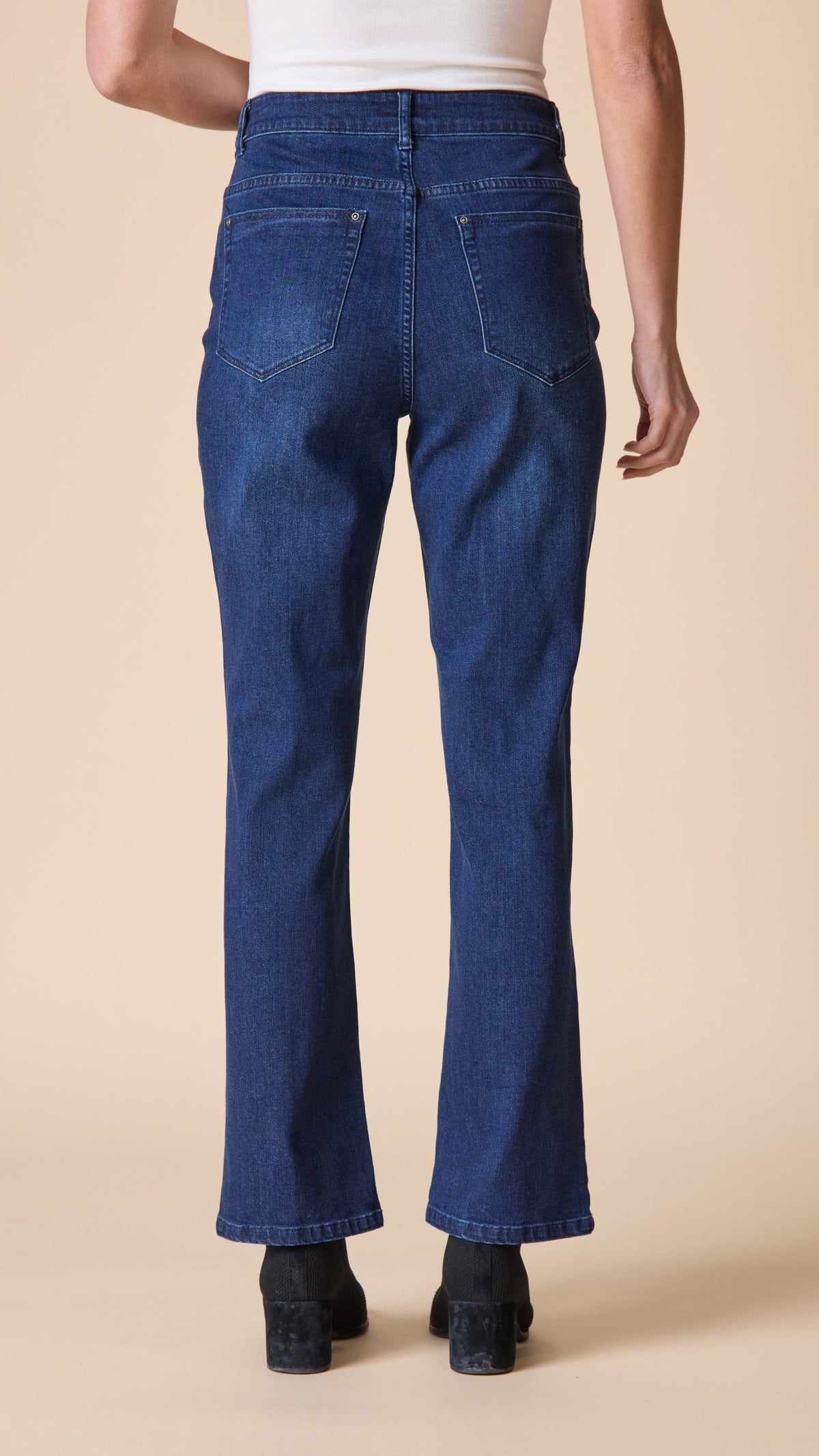 Back view of a person wearing dark blue straight-leg jeans with a white top and black shoes, standing against a solid beige background.