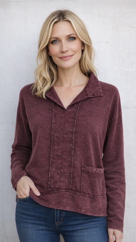 A woman with blonde hair wearing a maroon long-sleeve collared top with vertical button details and a front pocket, paired with blue jeans, standing against a light gray textured wall.