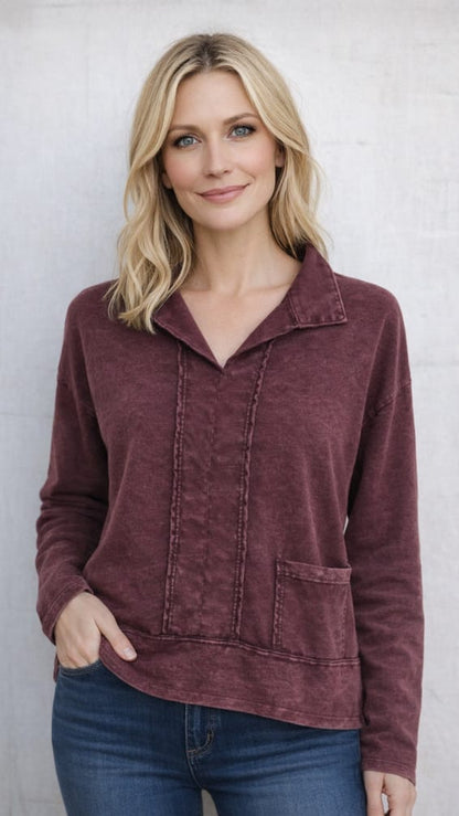 A woman with blonde hair wearing a maroon long-sleeve collared top with vertical button details and a front pocket, paired with blue jeans, standing against a light gray textured wall.