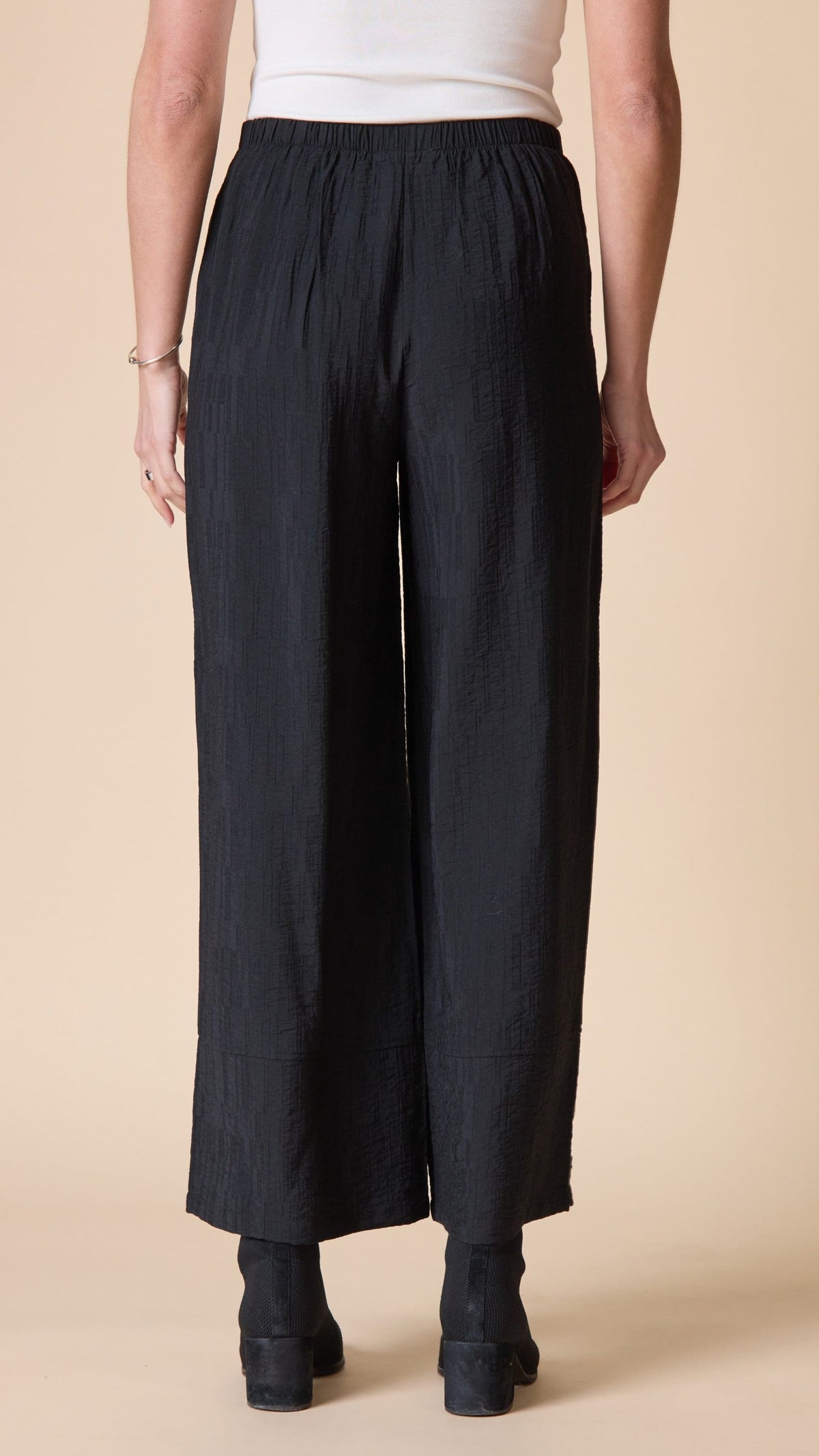 A person shown from the back wearing loose black wide-leg cropped pants with an elastic waistband, paired with a white top and black ankle boots, against a plain beige studio background.