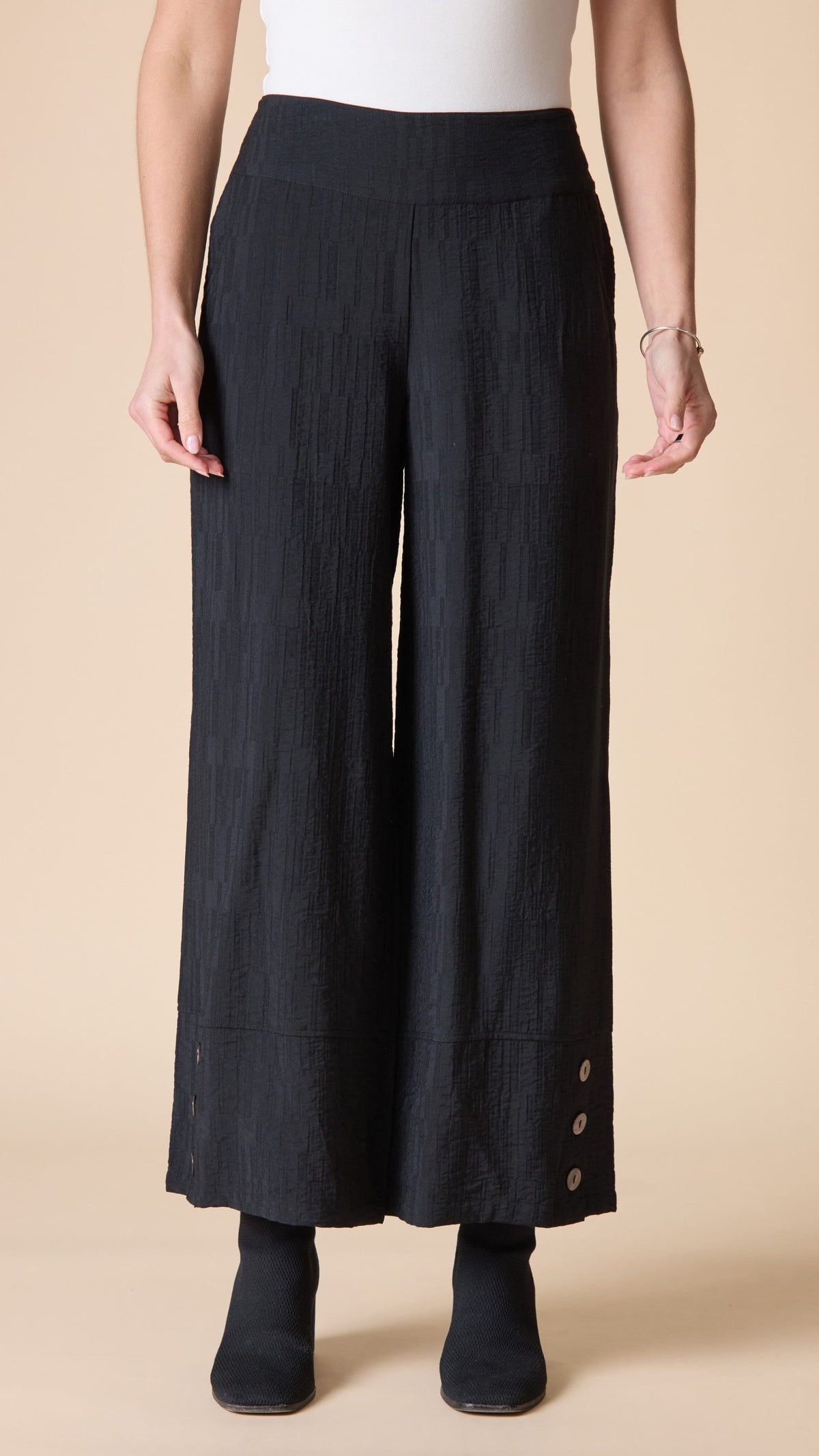 A model wearing black wide-leg cropped pants with a subtle vertical crinkle texture and decorative buttons on the lower hems, paired with a white top and black ankle boots against a solid beige background.