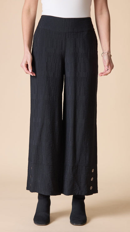 A model wearing black wide-leg cropped pants with a subtle vertical crinkle texture and decorative buttons on the lower hems, paired with a white top and black ankle boots against a solid beige background.