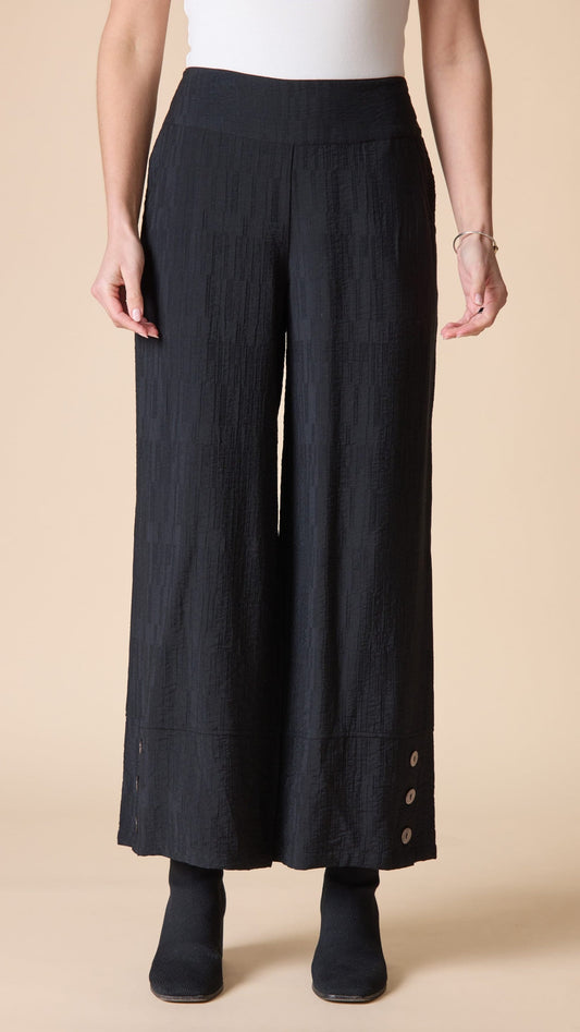 A model wearing black wide-leg cropped pants with a subtle vertical crinkle texture and decorative buttons on the lower hems, paired with a white top and black ankle boots against a solid beige background.