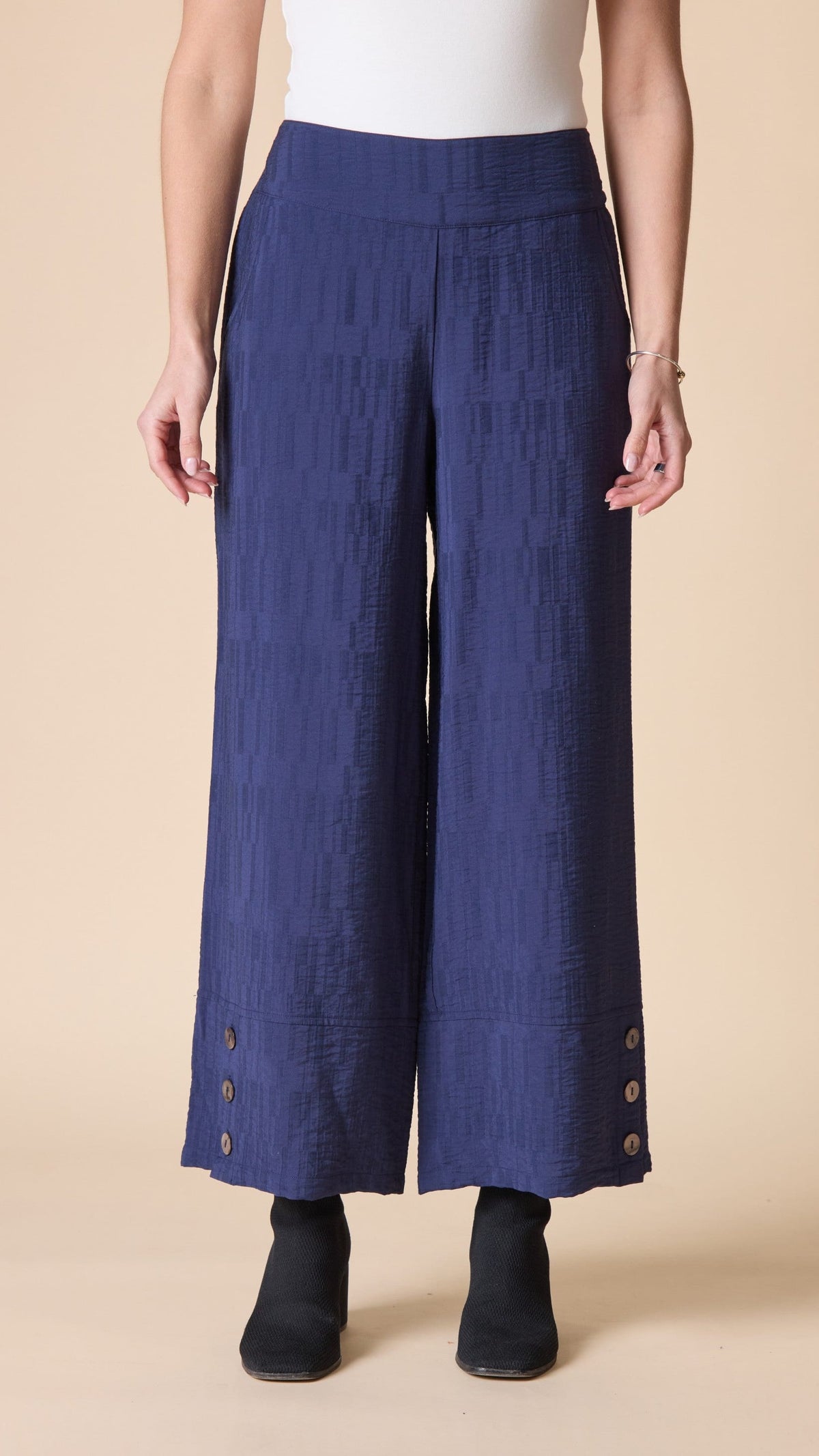 A model is shown from the waist down wearing textured, wide-leg cropped pants in a deep blue color, styled with a white top and black ankle boots against a solid beige background.