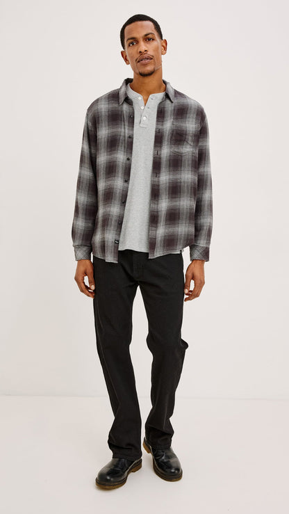 A man modeling a long-sleeve gray and black plaid button-down shirt over a white henley t-shirt, paired with black pants and black shoes, against a plain white studio background.