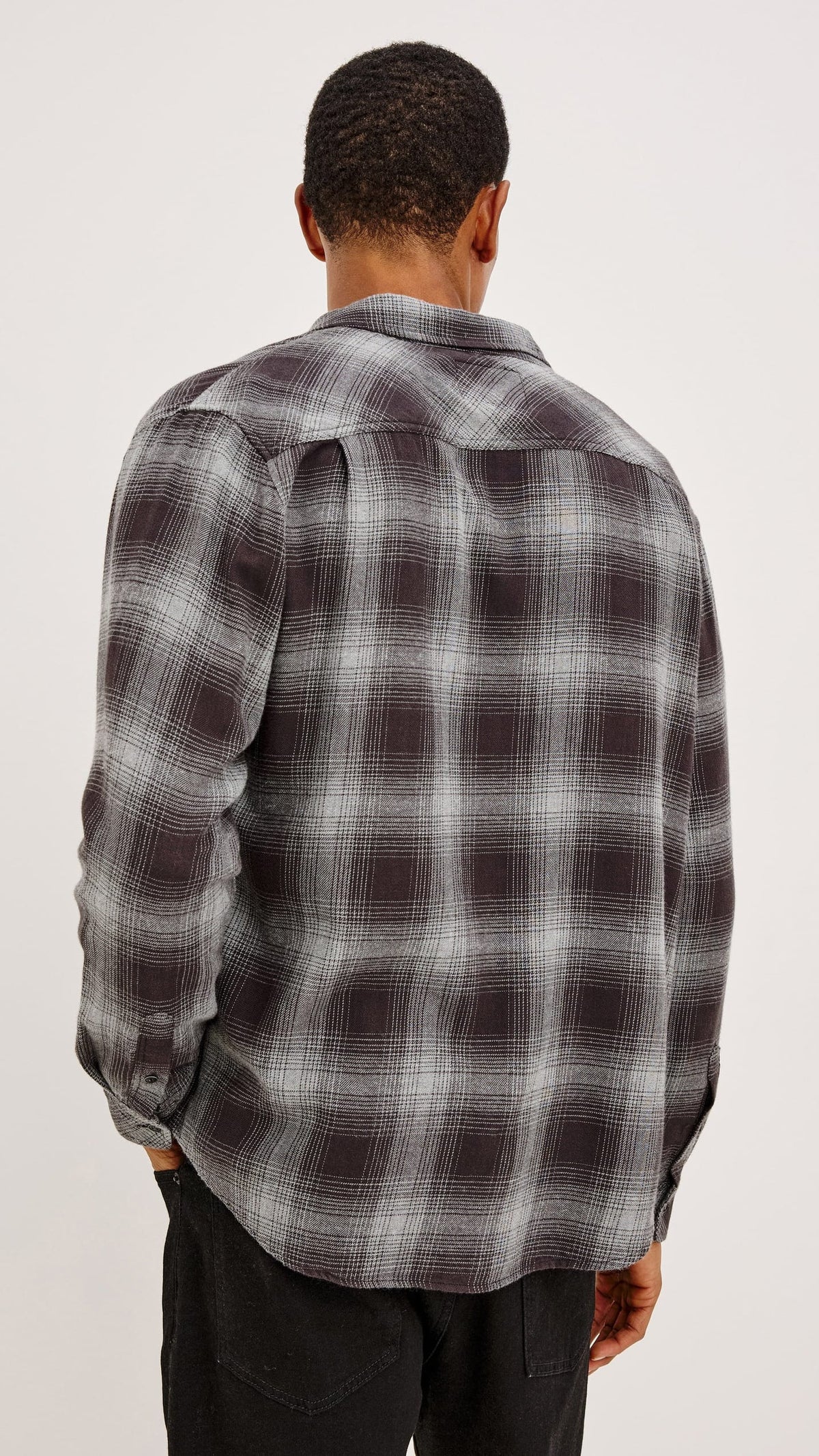 A man viewed from behind wearing a dark gray and black plaid flannel shirt with white grid lines, paired with black pants, against a plain light gray background.