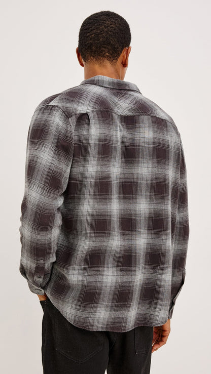 A man viewed from behind wearing a dark gray and black plaid flannel shirt with white grid lines, paired with black pants, against a plain light gray background.
