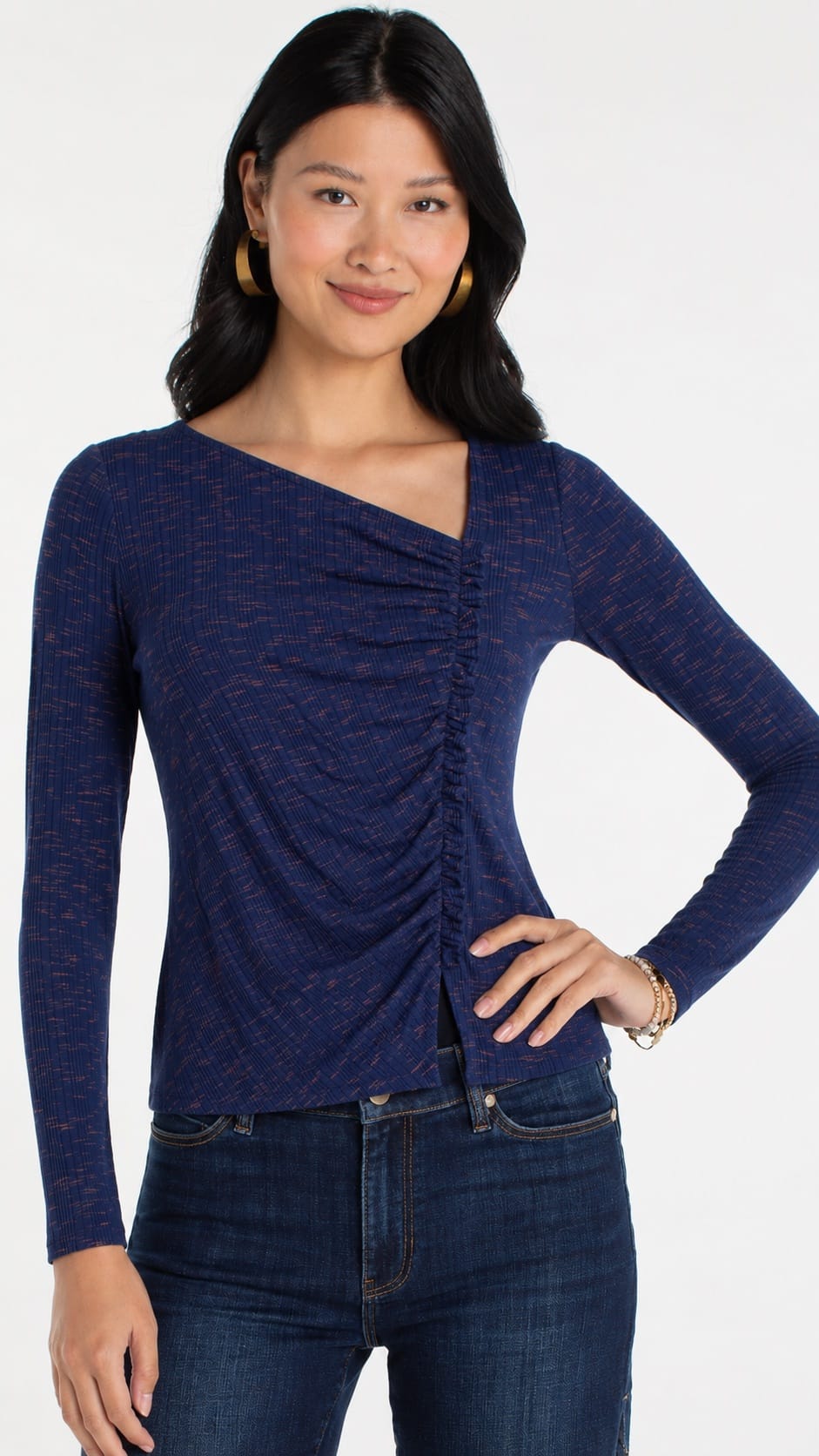 A woman wearing a navy blue long-sleeve top with a ruched front and asymmetrical neckline, paired with dark blue jeans, against a plain light gray background.