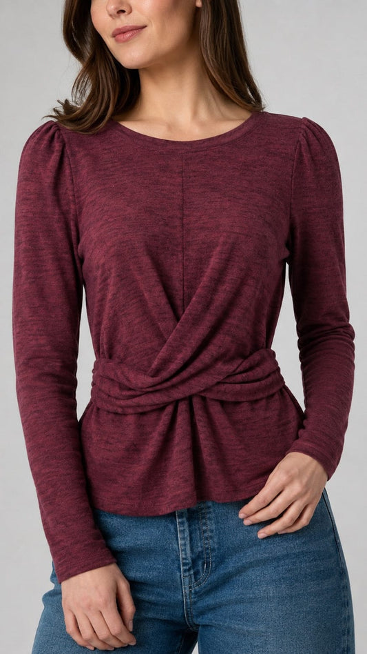 A woman wearing a heathered burgundy long-sleeve top with a twisted wrap-front detail at the waist, paired with blue jeans, against a plain gray studio background.