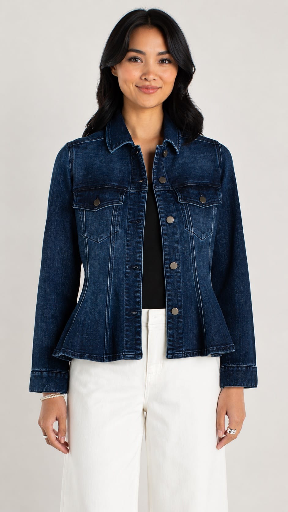 A woman modeling a dark blue denim jacket with a peplum waist, worn over a black top and white pants, against a light gray studio background.