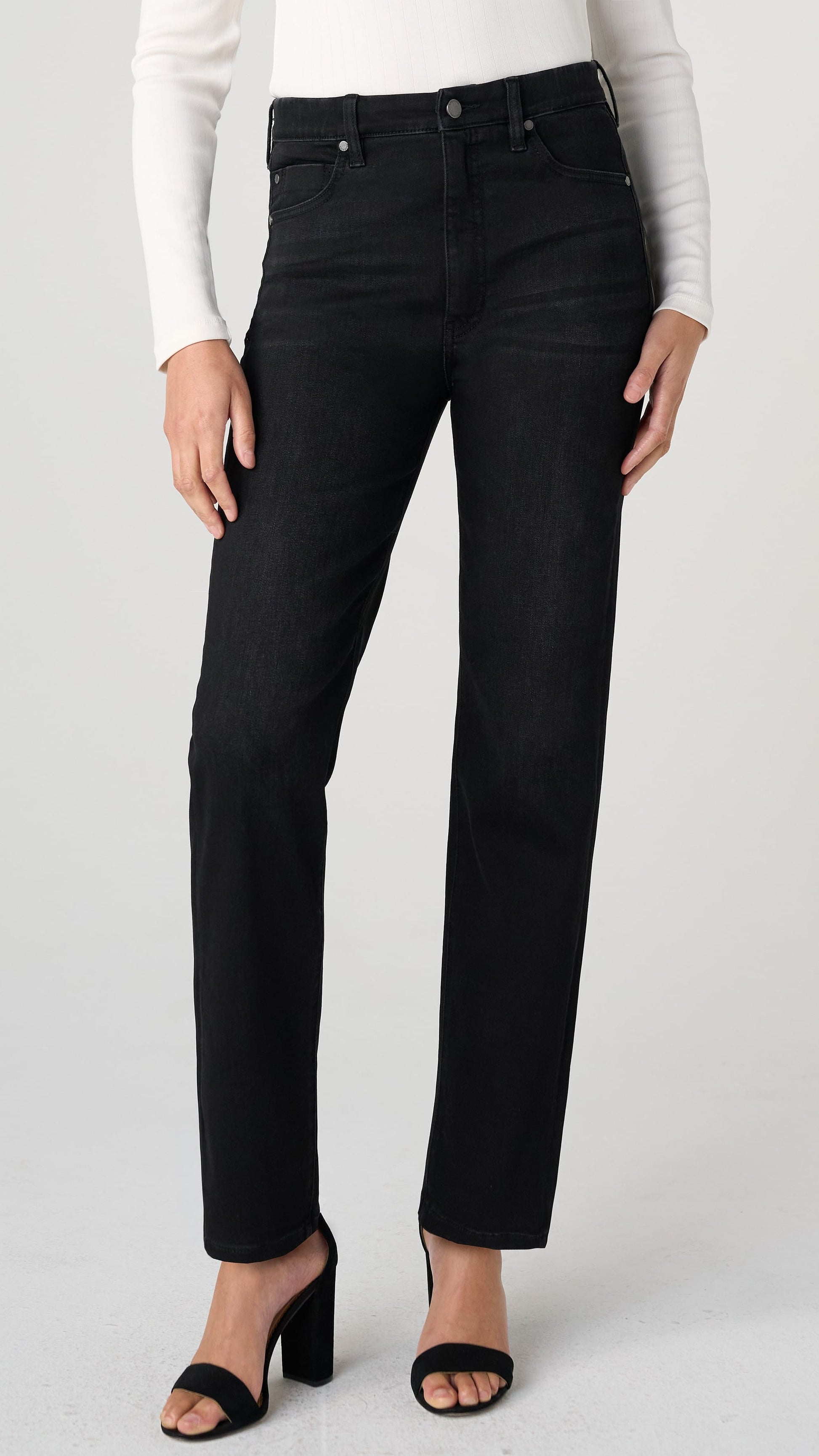 A model wearing a pair of black high-waisted straight-leg jeans, styled with a white long-sleeve top and black heeled sandals against a light gray studio background.