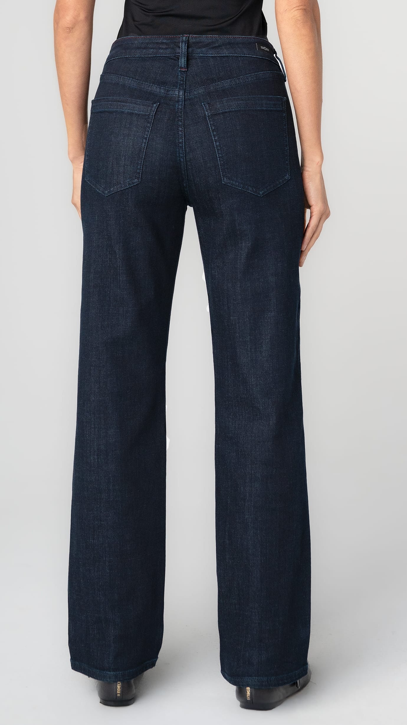 Rear view of a model wearing dark indigo wide-leg jeans with a black top, standing against a light gray studio background.