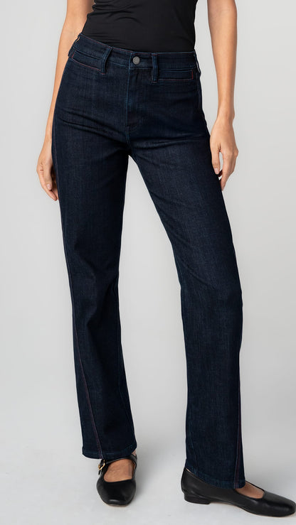 A model wearing a pair of dark navy blue straight-leg jeans with a subtle red contrast stitch, paired with a black top and black flats, against a light gray studio background.