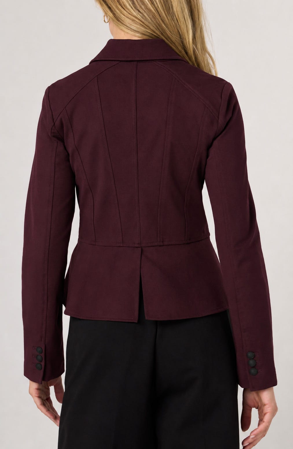 A woman shown from the back wearing a structured maroon blazer with a peplum waist and black trousers, against a light gray studio background.