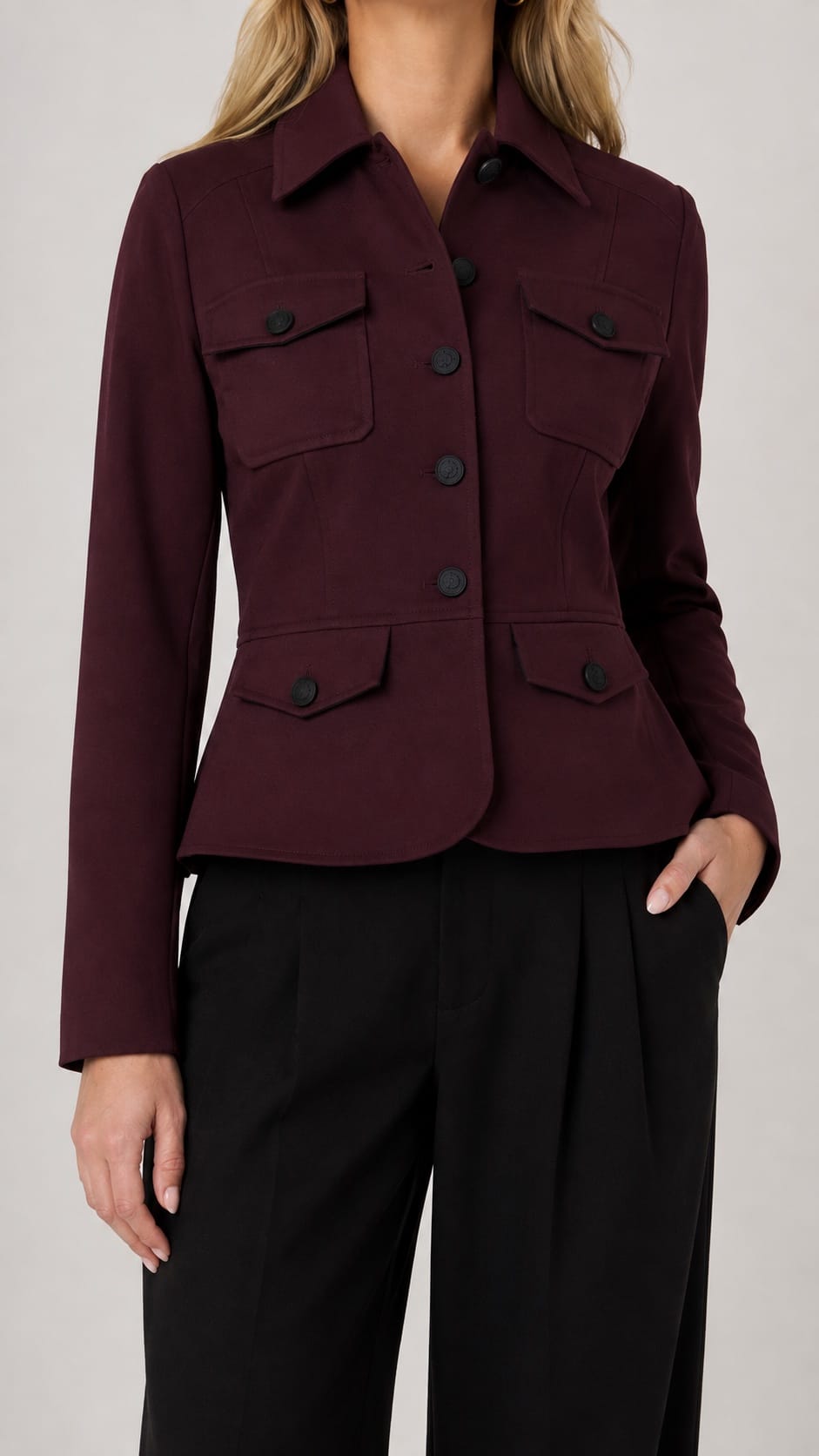 A woman wearing a cropped, long-sleeve burgundy jacket with four front flap pockets and black wide-leg trousers, shown from the neck down against a light gray studio background.