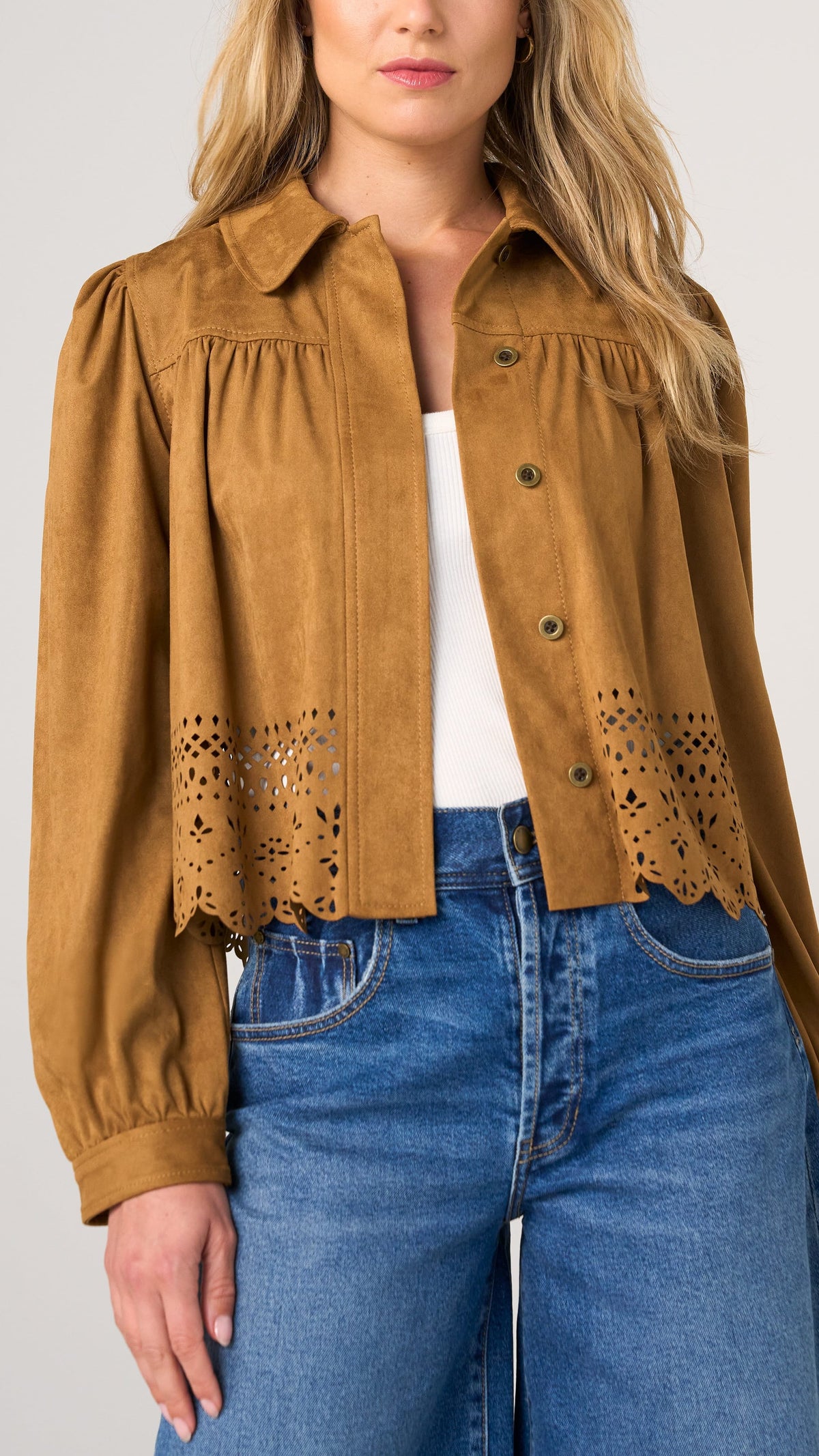 A woman wearing a cropped tan suede-like button-up shirt with eyelet lace detailing at the hem, styled over a white top and blue jeans against a light gray studio background.