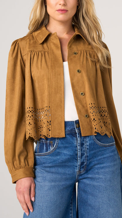 A woman wearing a cropped tan suede-like button-up shirt with eyelet lace detailing at the hem, styled over a white top and blue jeans against a light gray studio background.