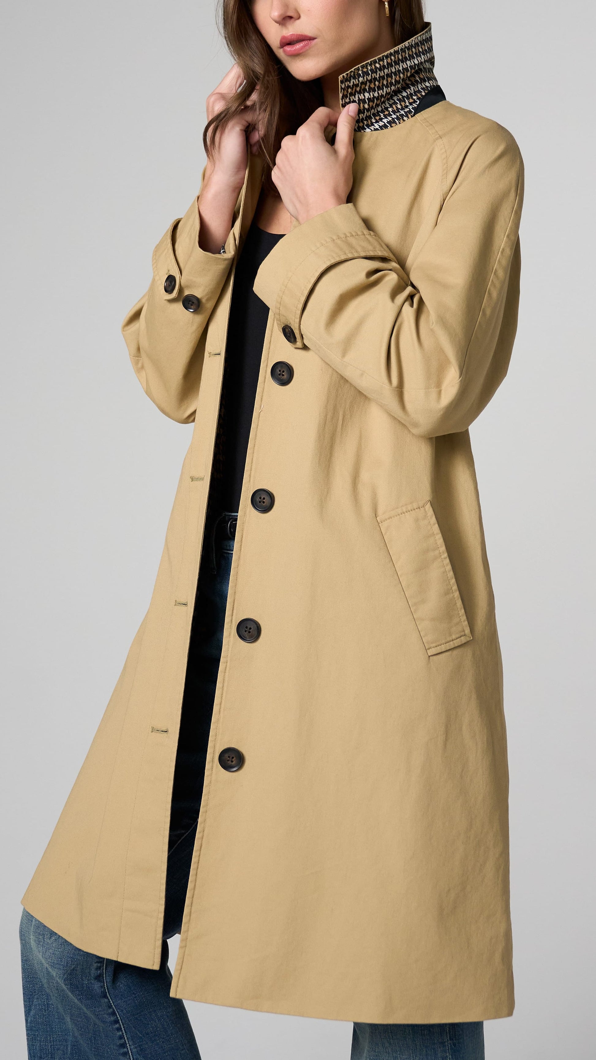 A woman wearing a beige trench coat over a black top and blue jeans, shown from the shoulders to the upper thighs against a light gray studio background.