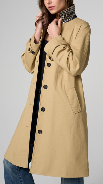 A woman wearing a beige trench coat over a black top and blue jeans, shown from the shoulders to the upper thighs against a light gray studio background.