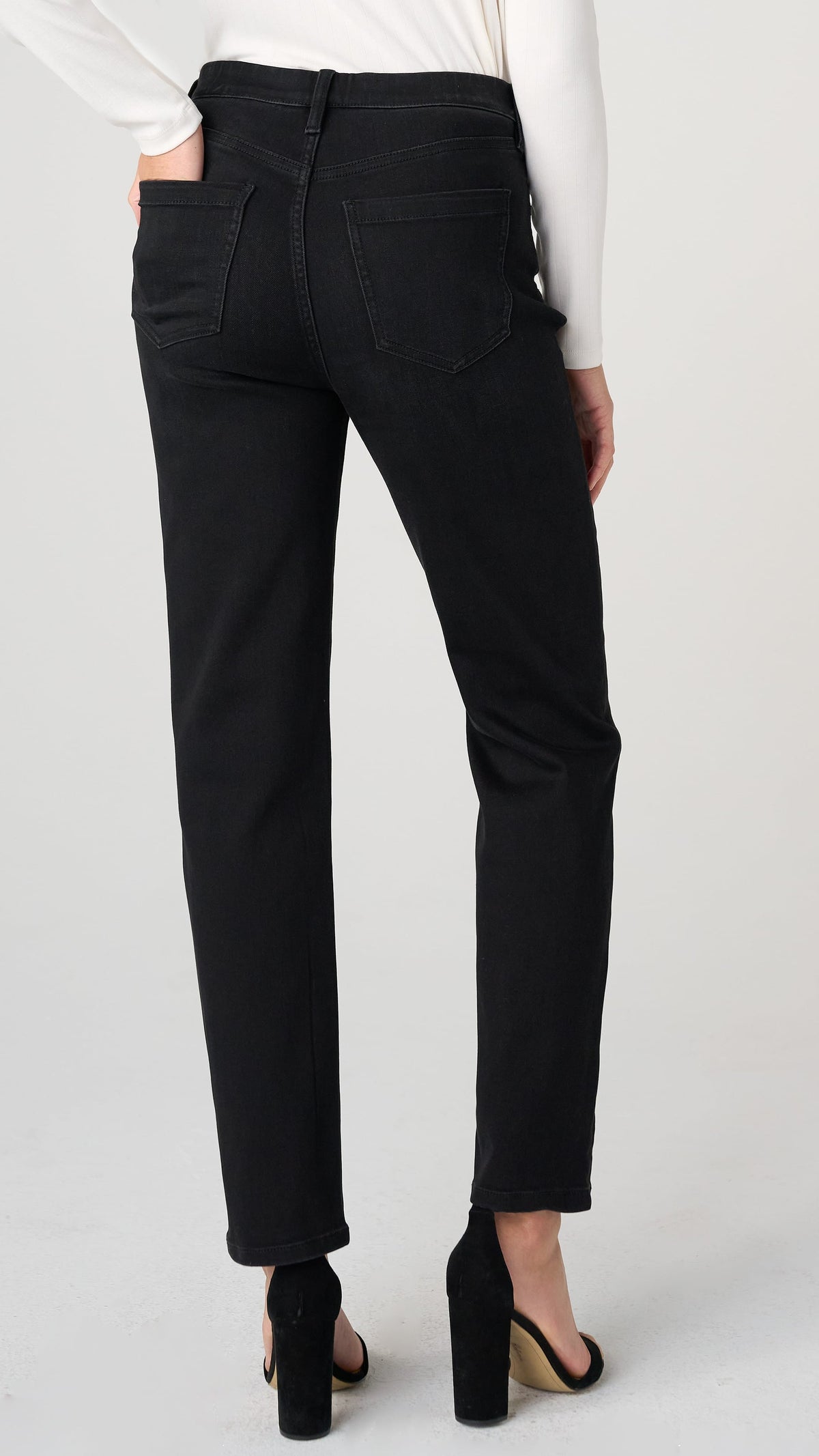 Rear view of a model wearing black slim-fit jeans with a white long-sleeve top and black high heels, set against a plain light gray studio background.