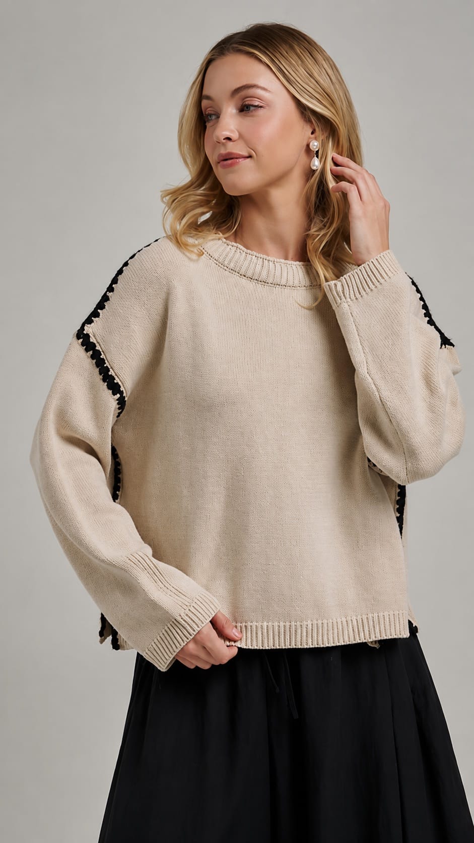 A woman wearing a cream-colored knit sweater with black tassel trim along the shoulders and sleeves, paired with a black skirt, against a neutral gray studio background.