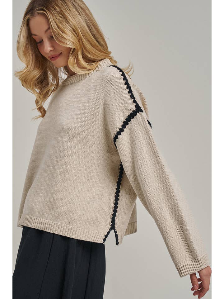 A woman wearing a beige knit sweater with black braided trim along the shoulder seams, paired with dark pleated trousers, photographed against a plain light gray studio background.