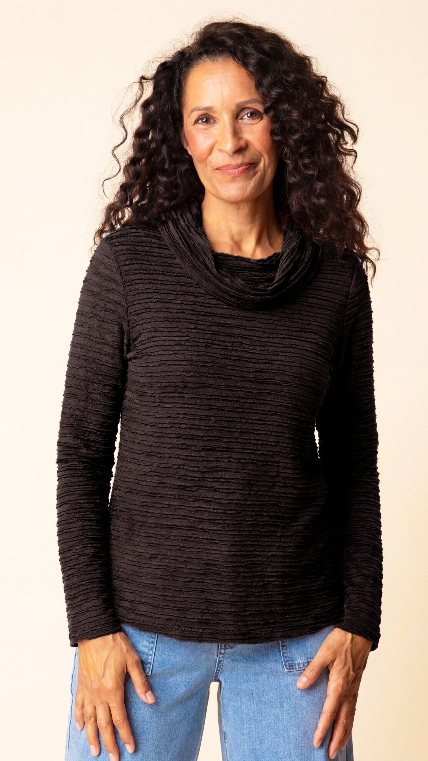 A woman with long, curly dark hair wearing a dark brown ribbed cowl-neck long-sleeve top and light blue jeans, standing against a plain beige background.