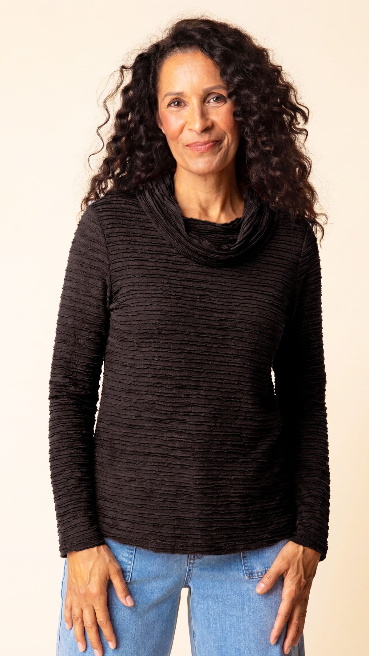 A woman with long, curly dark hair wearing a dark brown ribbed cowl-neck long-sleeve top and light blue jeans, standing against a plain beige background.