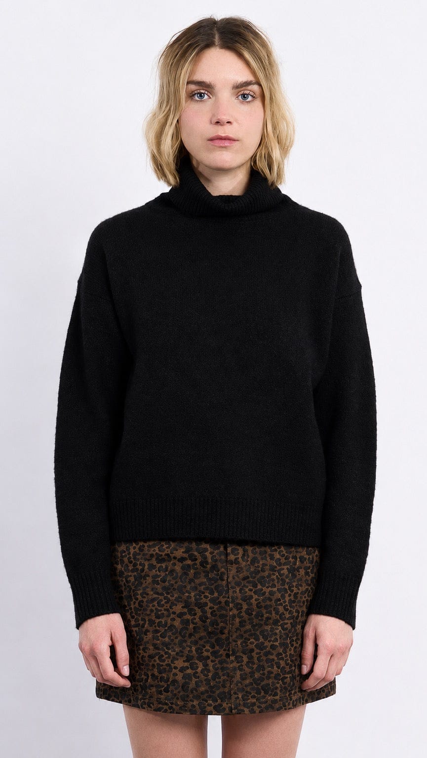 A woman wearing a black knit turtleneck sweater and a brown leopard-print mini skirt, standing against a light gray studio background.