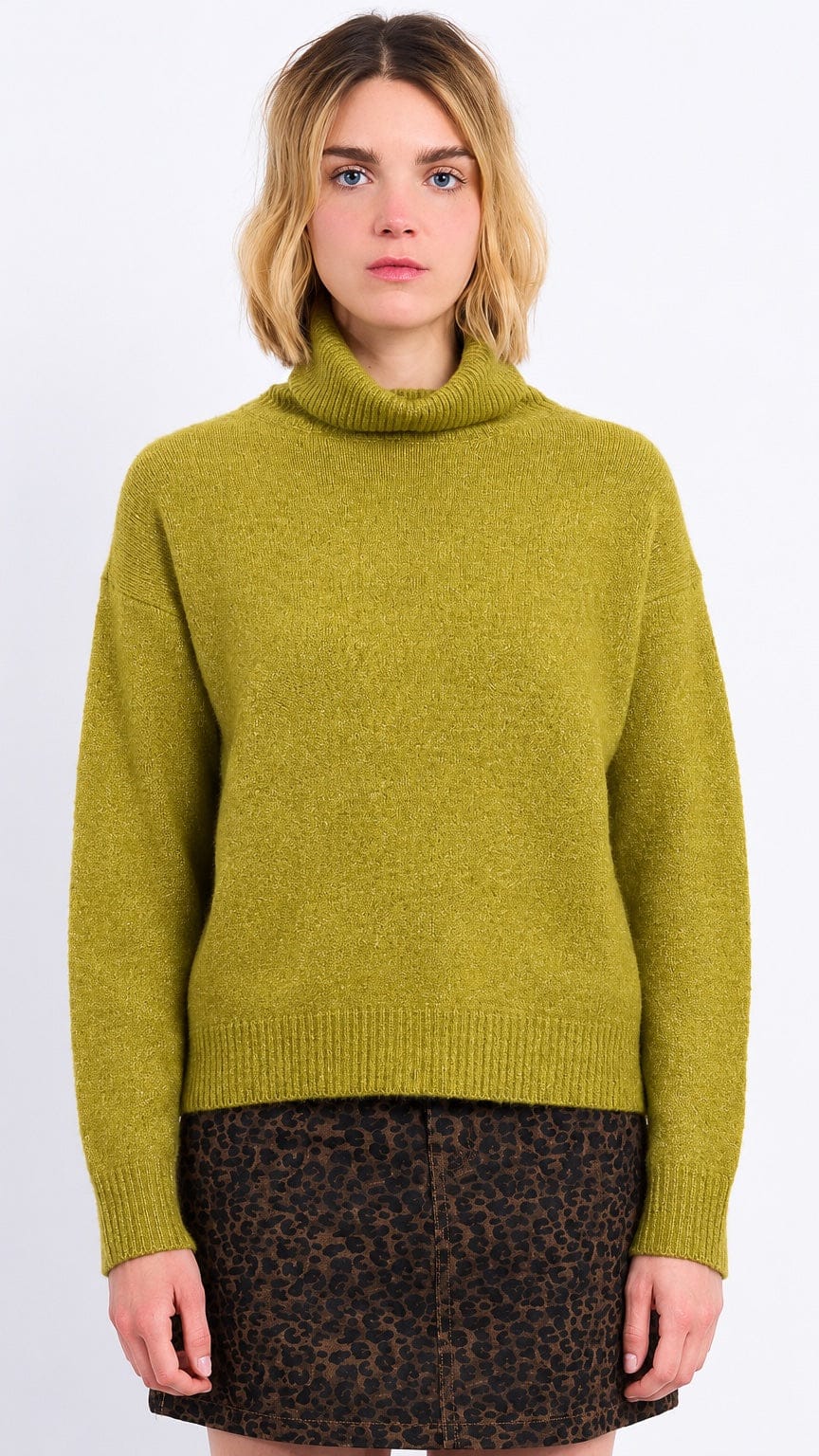 A woman wearing a fuzzy olive green turtleneck sweater and a brown leopard-print mini skirt, standing against a plain white studio background.