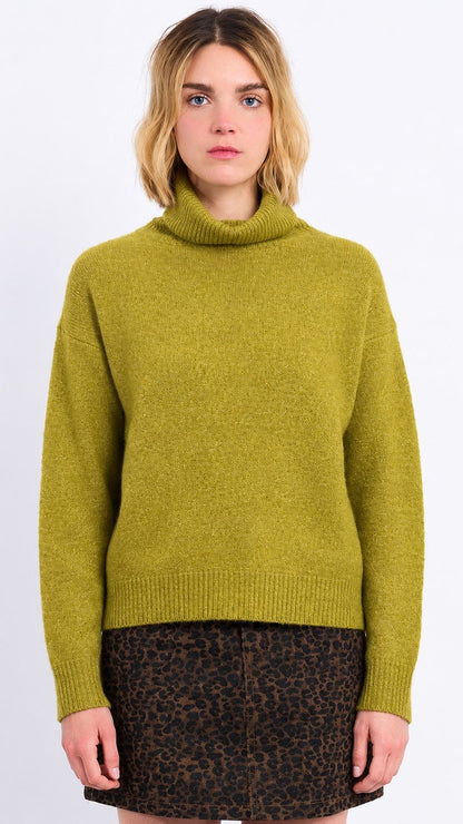 A woman wearing a fuzzy olive green turtleneck sweater and a brown leopard-print mini skirt, standing against a plain white studio background.