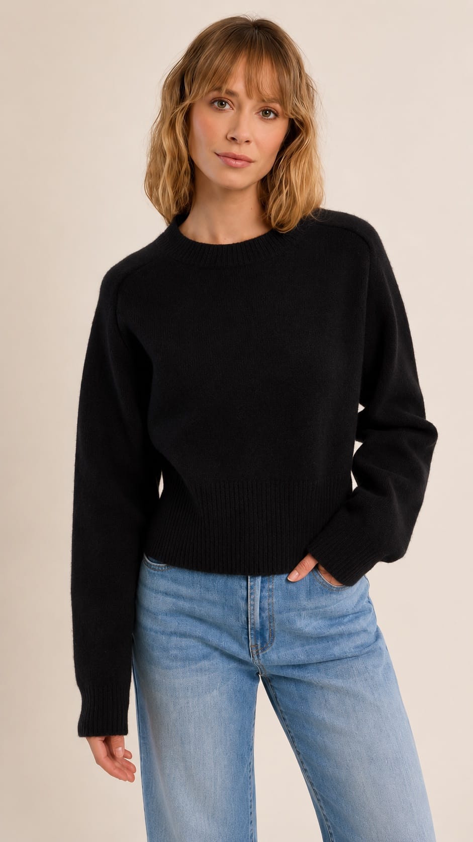A woman wearing a black long-sleeve knit sweater and light blue jeans, standing against a plain beige studio background.