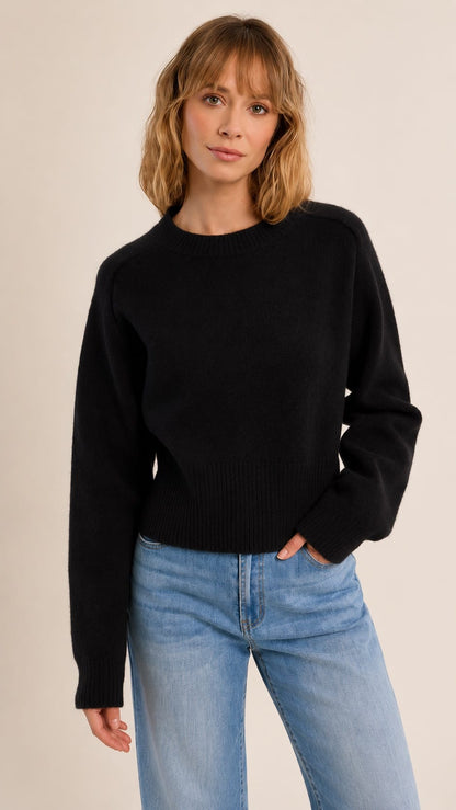 A woman wearing a black long-sleeve knit sweater and light blue jeans, standing against a plain beige studio background.