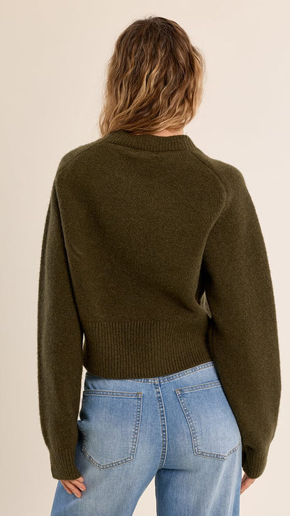 A woman viewed from the back wearing an olive green knit sweater and light blue jeans against a plain beige studio background.