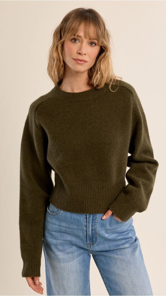 A woman wearing a dark olive green knit crewneck sweater and light blue jeans, standing against a plain beige studio background.