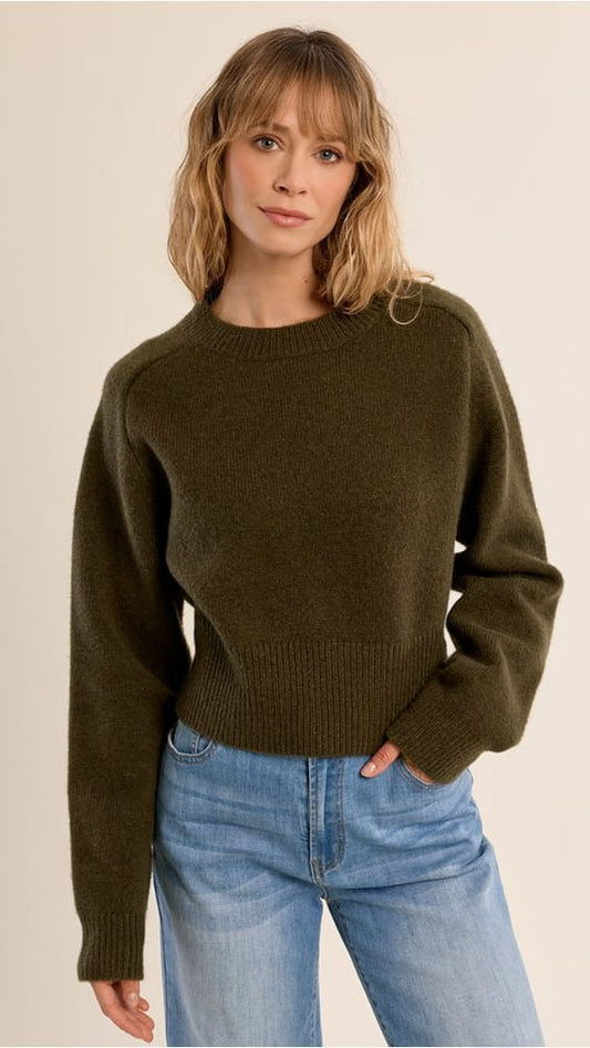 A woman wearing a dark olive green knit crewneck sweater and light blue jeans, standing against a plain beige studio background.