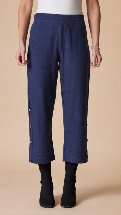 A model is shown from the waist down wearing navy blue cropped trousers with side buttons, paired with black ankle boots against a solid beige background.