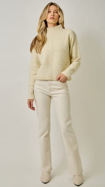 A woman modeling a cream-colored knit sweater and matching cream pants against a plain light gray studio background.