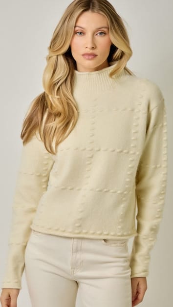 A woman wearing a cream-colored knit mock neck sweater with textured raised patterns, paired with cream pants, against a light gray studio background.