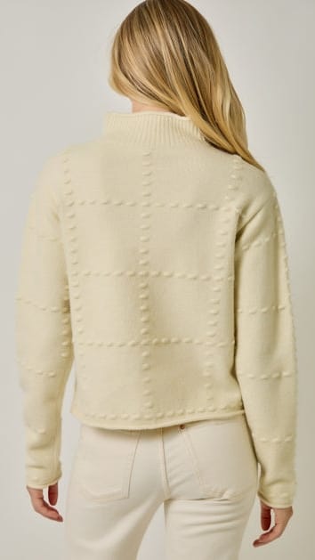A woman viewed from the back wearing a cream-colored knit sweater with a textured grid pattern and a mock neck, paired with cream pants, against a light gray studio background.