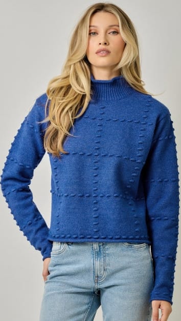 A woman wearing a textured royal blue mock neck sweater with raised knit details and light-wash blue jeans, photographed against a neutral light gray studio background.