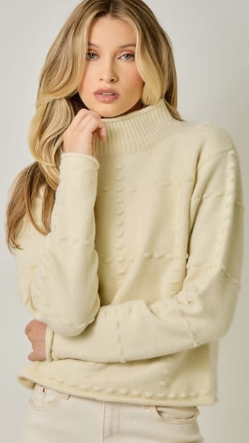 A woman wearing a cream-colored ribbed turtleneck sweater with textured knit details on the front, paired with light beige pants, set against a plain light gray studio background.