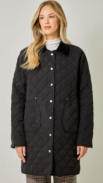 A blonde woman wearing a black diamond-quilted coat with a dark collar and white buttons, standing against a light gray studio background.