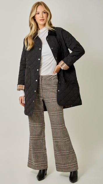 A woman models a black quilted jacket over a white top and brown plaid flared trousers, standing against a neutral light gray studio background.