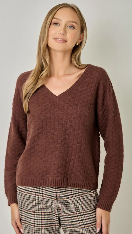 A woman wearing a brown ribbed knit V-neck sweater and plaid pants, standing against a plain light gray studio background.