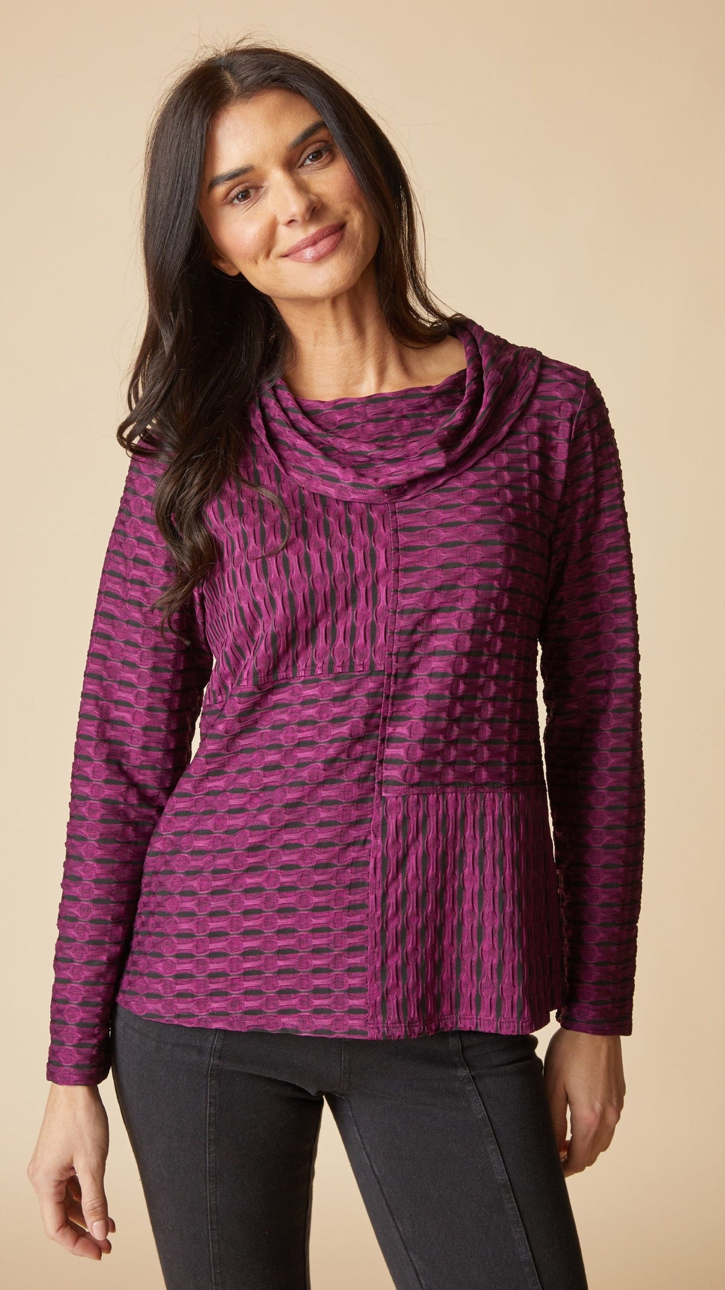A woman wearing a textured, long-sleeve cowl-neck top in a purple and black patterned fabric, paired with black pants, against a solid beige background.