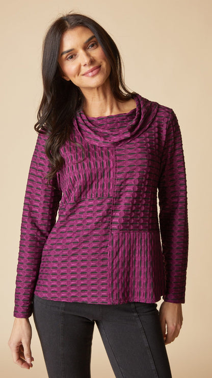 A woman wearing a textured, long-sleeve cowl-neck top in a purple and black patterned fabric, paired with black pants, against a solid beige background.