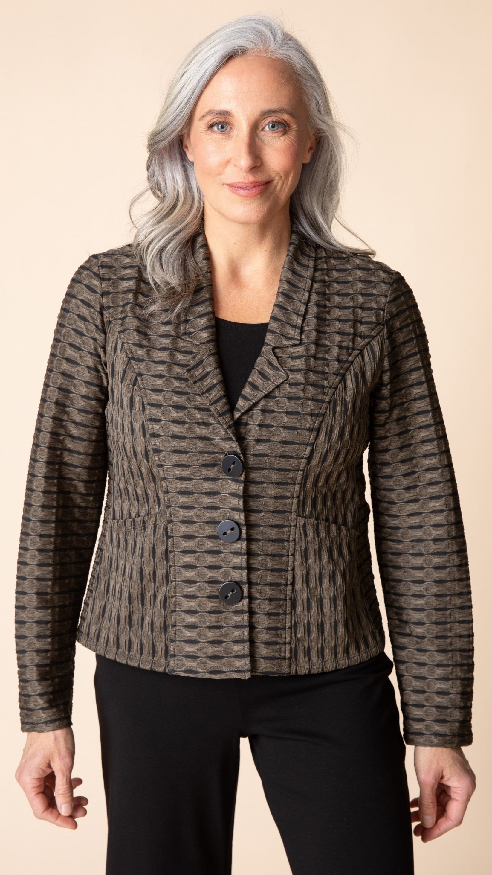 A woman with gray hair wearing a textured, patterned brown and gray blazer over a black top and black pants, standing against a solid beige background.
