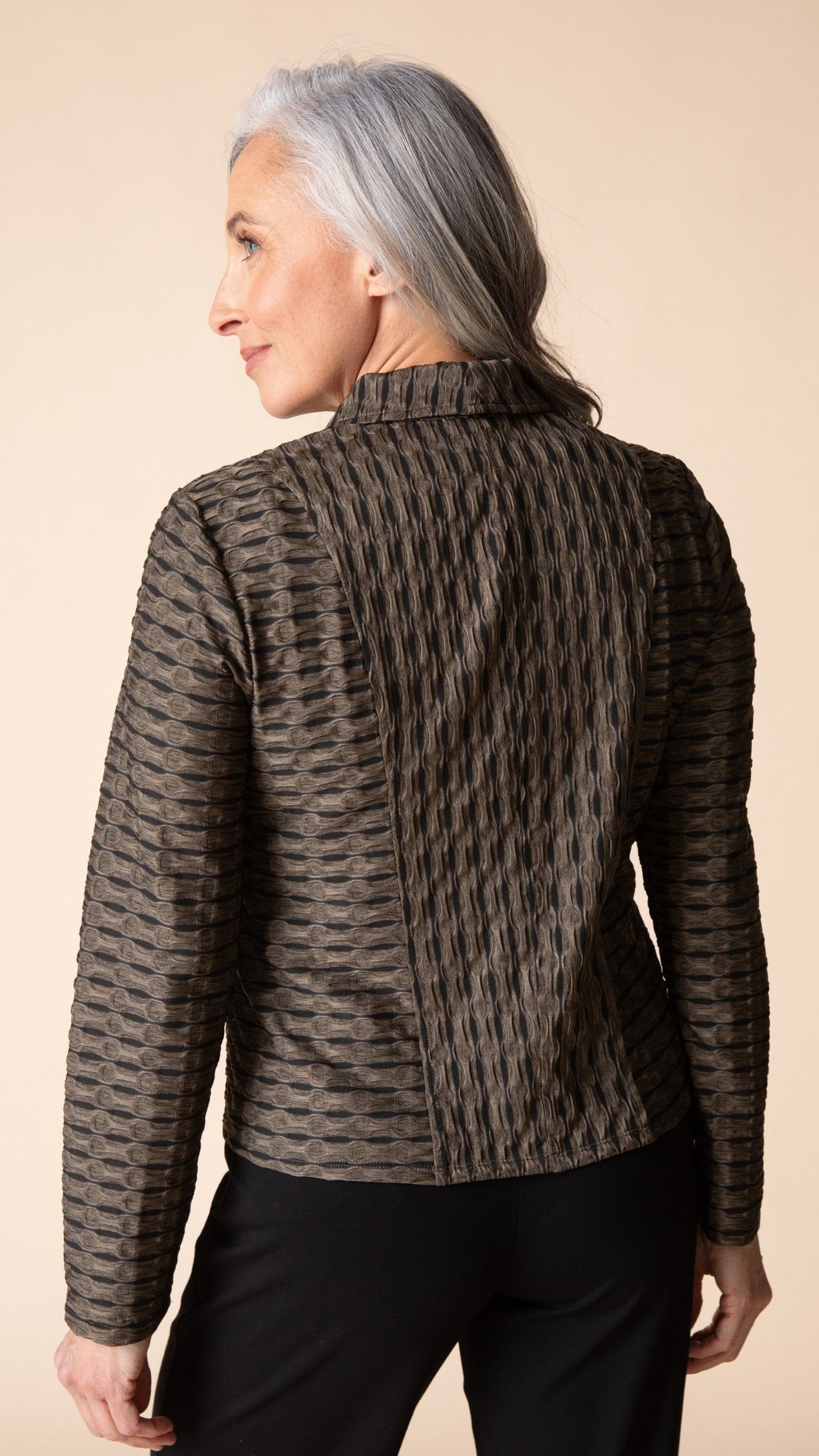 A woman with gray hair is shown from the back wearing a textured, patterned brown and black jacket with black pants, set against a solid beige background.