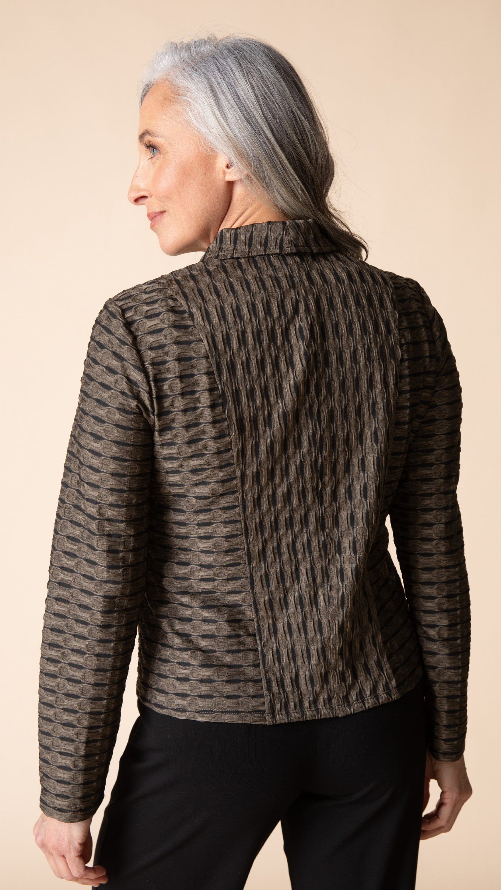 A woman with gray hair is shown from the back wearing a textured, patterned brown and black jacket with black pants, set against a solid beige background.